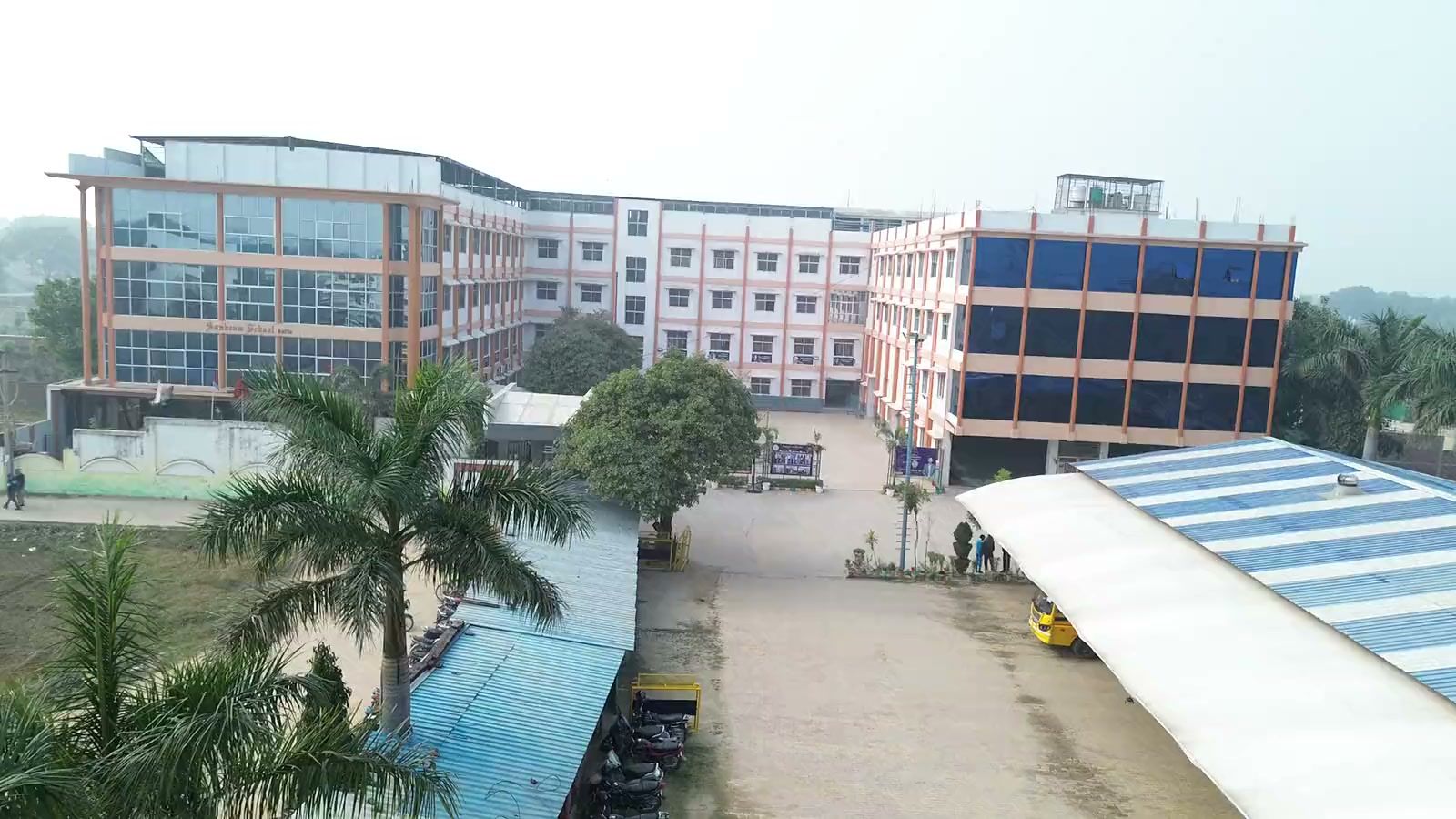 Sunbeam School Ballia from the air: the main teaching block with its glass frontage, the school name along the façade, and the drive and covered cycle stands running past it.